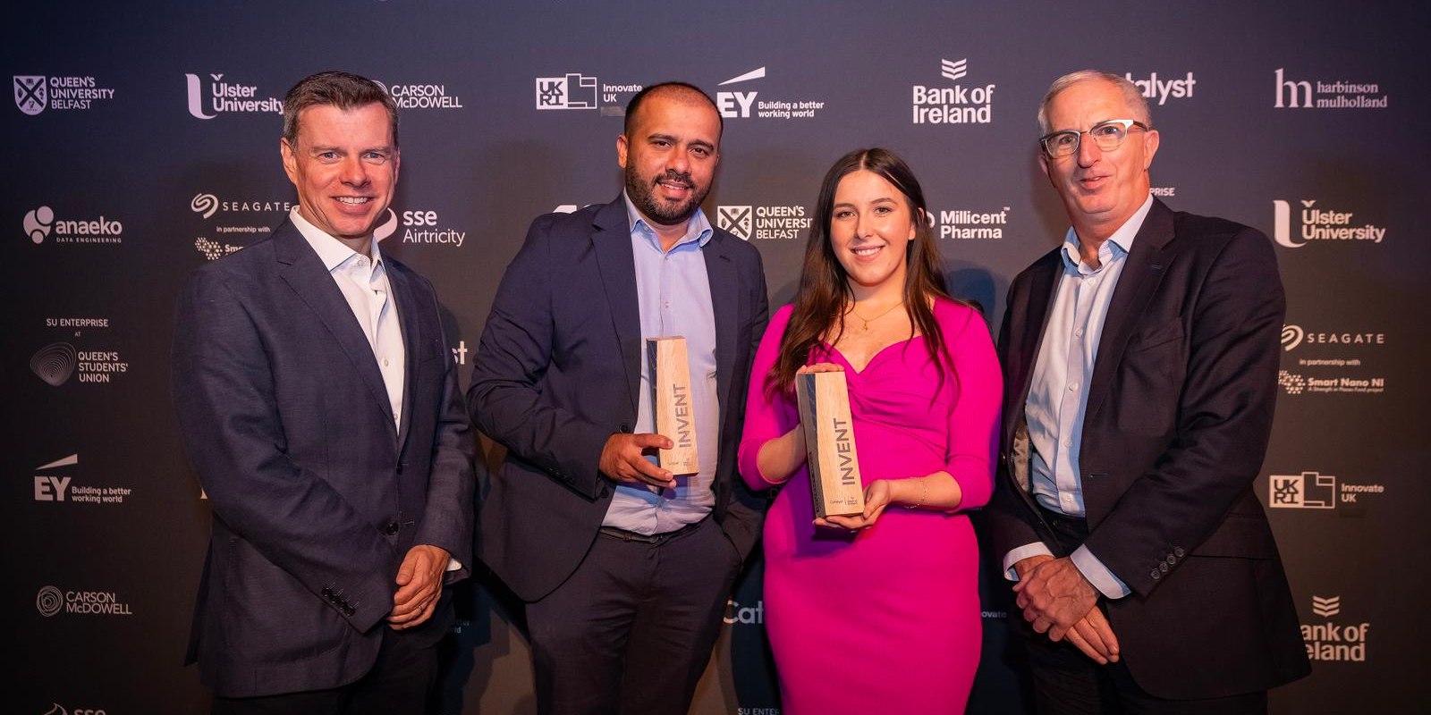 Andrine Mendez and GoPlugable co-founder Maebh accepting the Invent 23 award on stage, holding wooden trophies, alongside event organisers and a backdrop of sponsor and partner logos including Catalyst, Ulster University, Queen's University Belfast, EY, and Bank of Ireland